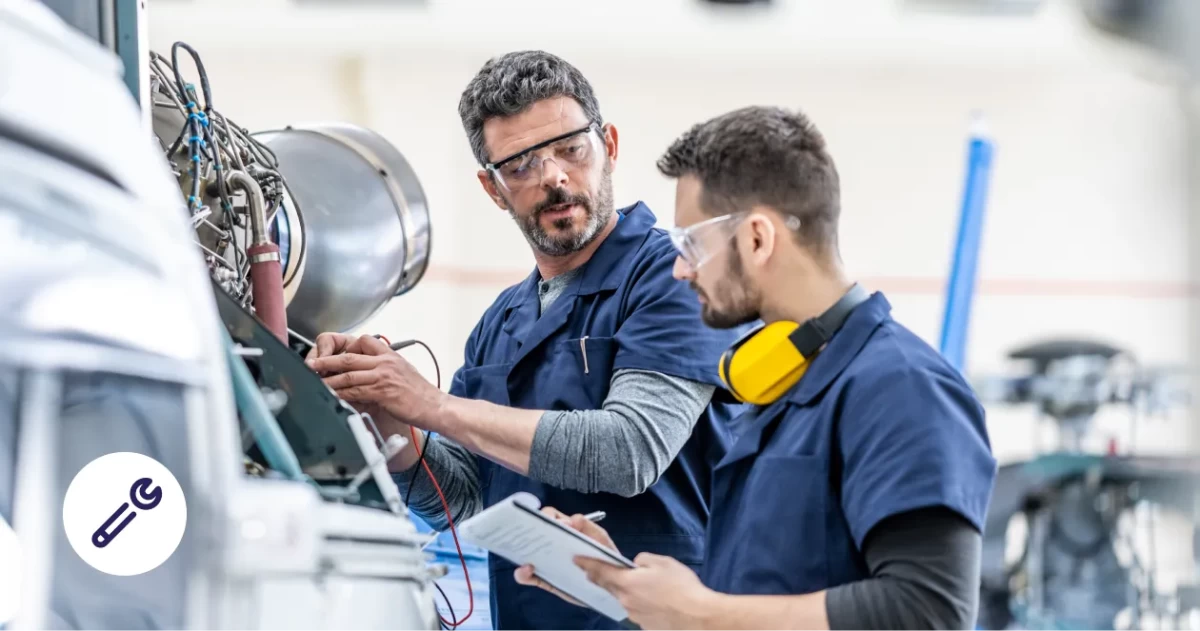 Zwei Mechaniker in blauer Arbeitskleidung tragen Schutzbrillen und arbeiten gemeinsam an einer komplexen Maschine in einer Werkstatt.