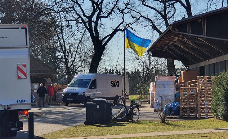 Zwei Lieferwagen und mehrere Menschen sind auf einem Hof mit einer Ukraine-Flagge zu sehen