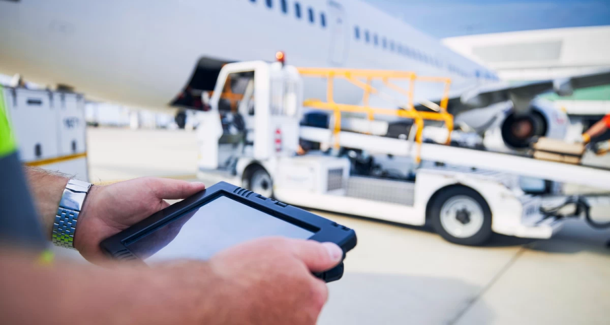 A worker uses a rugged tablet beside airport ground-support equipment and an aircraft