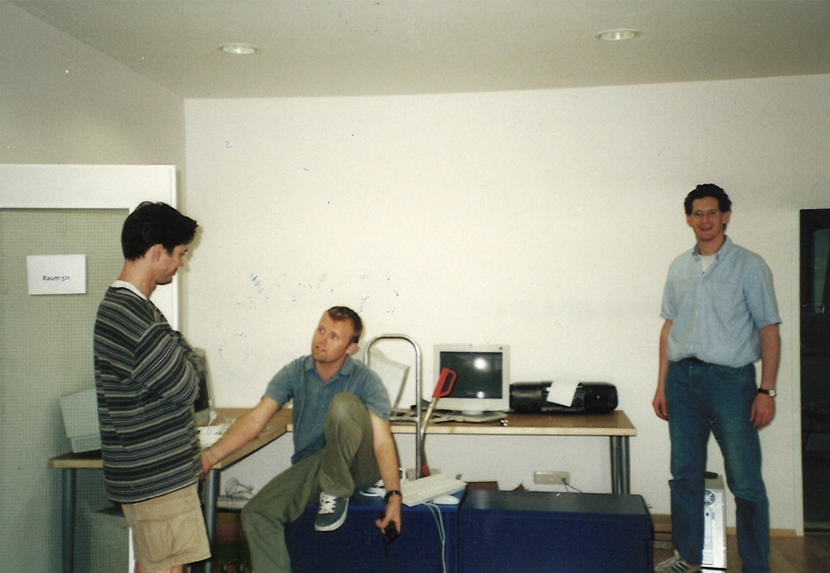 Three men in a room with a desk and a computer